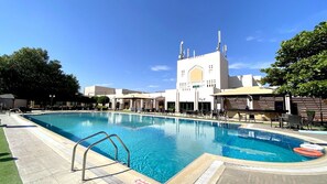 Une piscine dotée d’un poste de surveillance pour les sauveteurs, entourée de chaises longues et de parasols, avec un bâtiment en arrière-plan qui se distingue par une grande tour et des minarets.