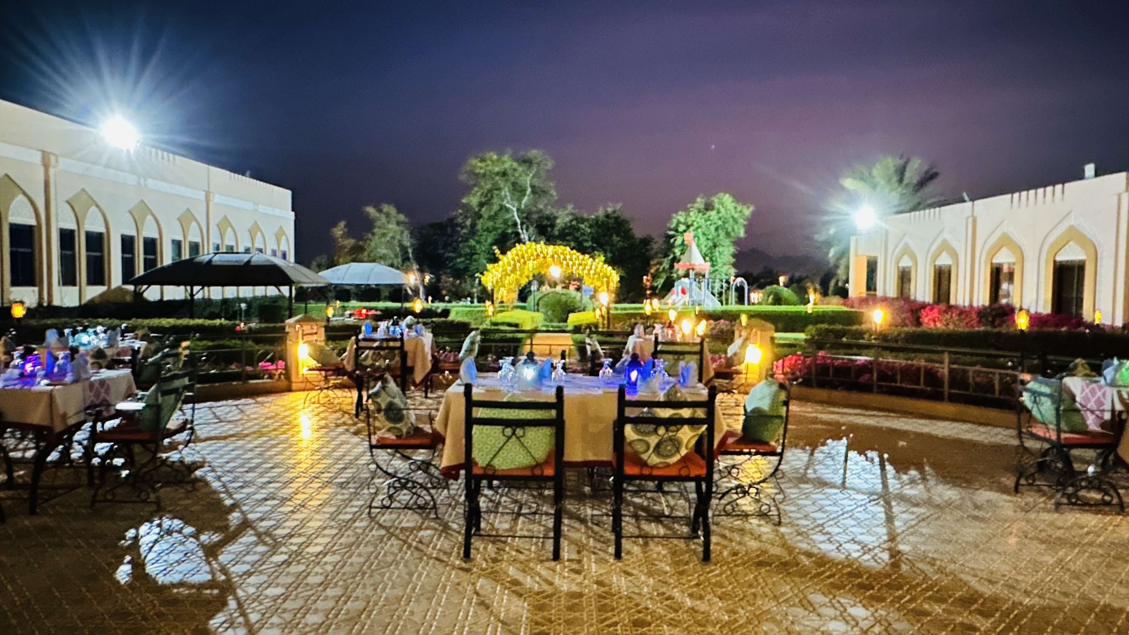 An outdoor dining area with tables and chairs, a fountain, and a well-lit garden at night.