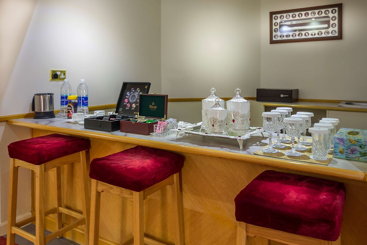 Zona de bar con taburetes, una cafetera, botellas de agua, un menú enmarcado y cristalería decorativa.