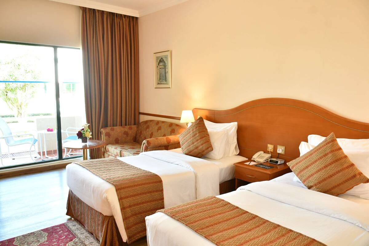 A hotel room with two beds, a small table, a chair, and a large window with curtains.
