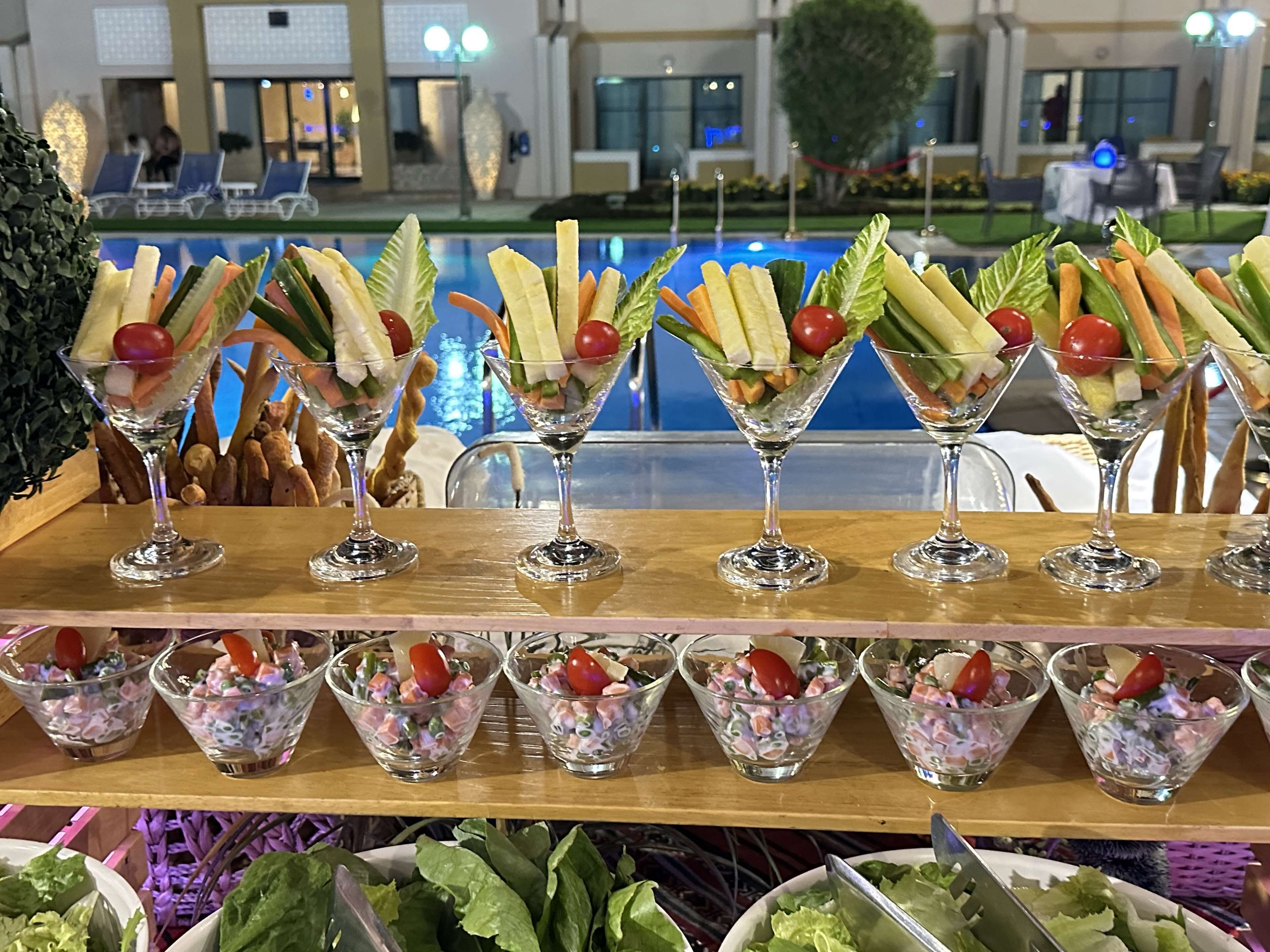 Mesa de buffet com diversos alimentos, com vista para a piscina ao fundo.