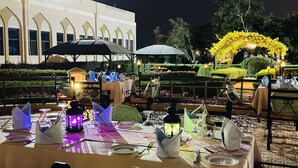 Un espace de restauration en plein air, avec des tables dressées pour un repas, entouré d’un jardin et d’un bâtiment doté de fenêtres en arc de cercle.