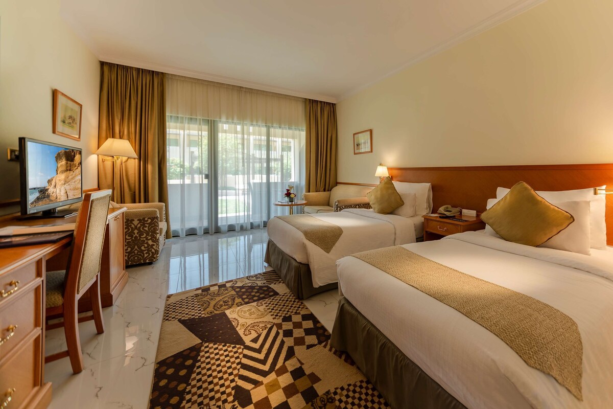 Une chambre d’hôtel avec deux lits, un bureau avec un ordinateur, une télévision et un tapis à motifs.