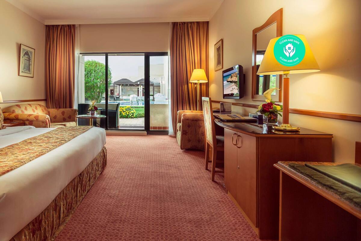 A hotel room with a large bed, a desk, a chair, and a television. There is a window with curtains and a view of the outside.