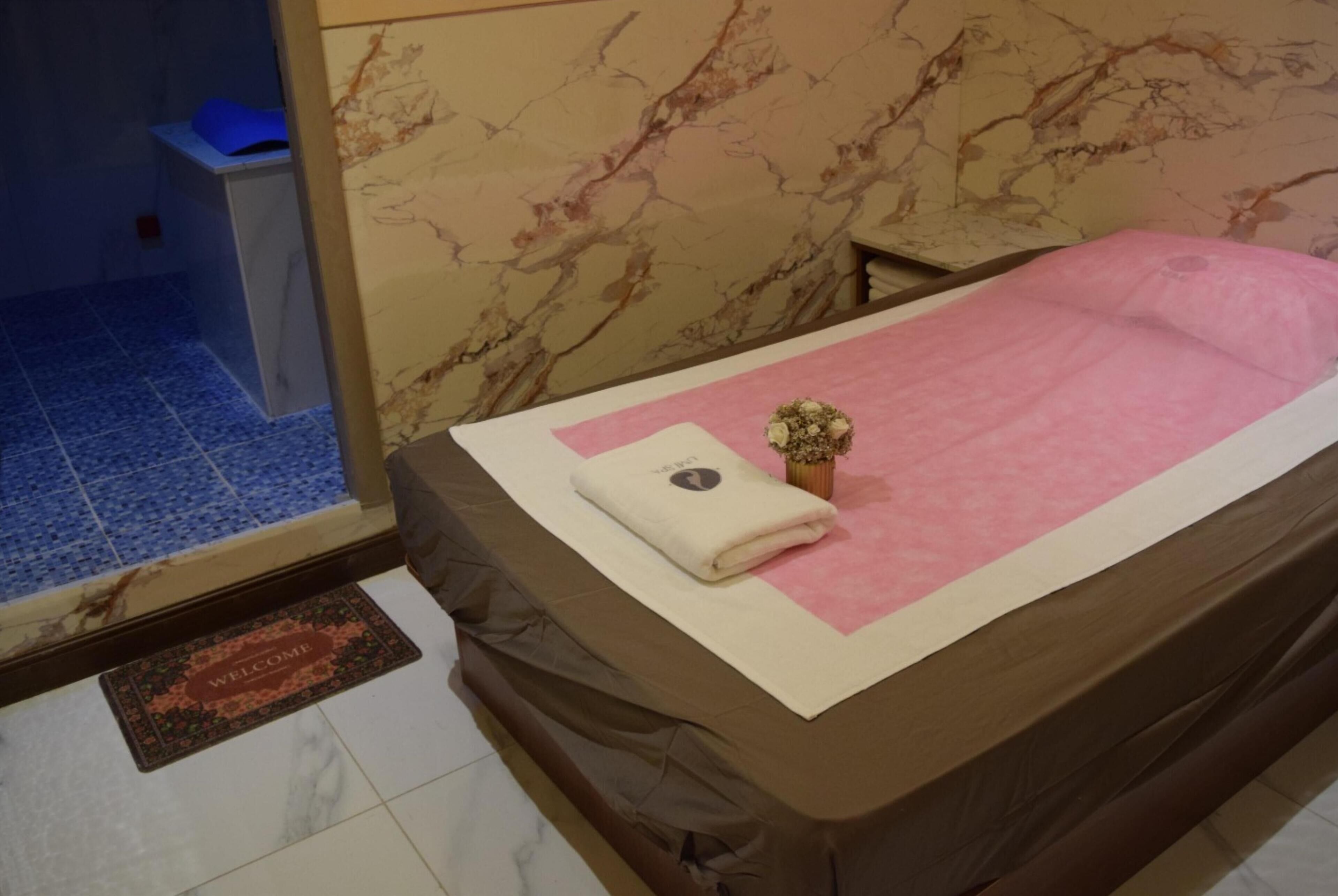 A room with a bed covered in a pink blanket, a small potted plant, and a welcome mat.