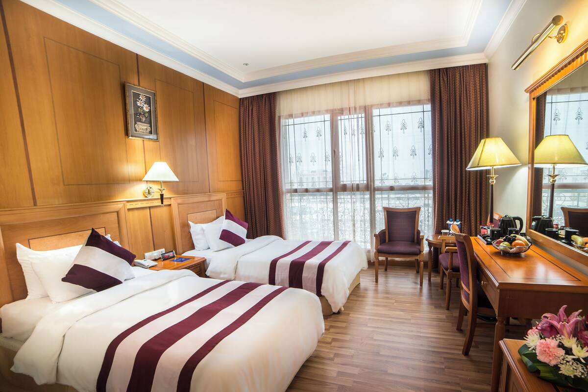 Een hotelkamer met twee bedden, een bureau, een stoel en een groot raam met gordijnen.