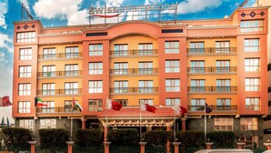 Um hotel com uma placa bem visível com a inscrição "Swiss International". Apresenta várias varandas com guarda-corpos, uma entrada central com um arco decorativo e diversas bandeiras hasteadas.