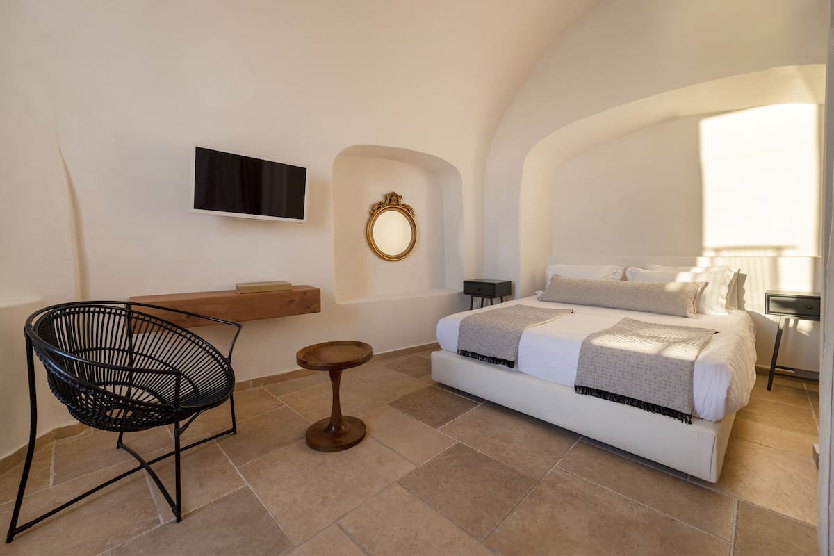 A bedroom with a bed, a chair, a small table, a TV, and a mirror.