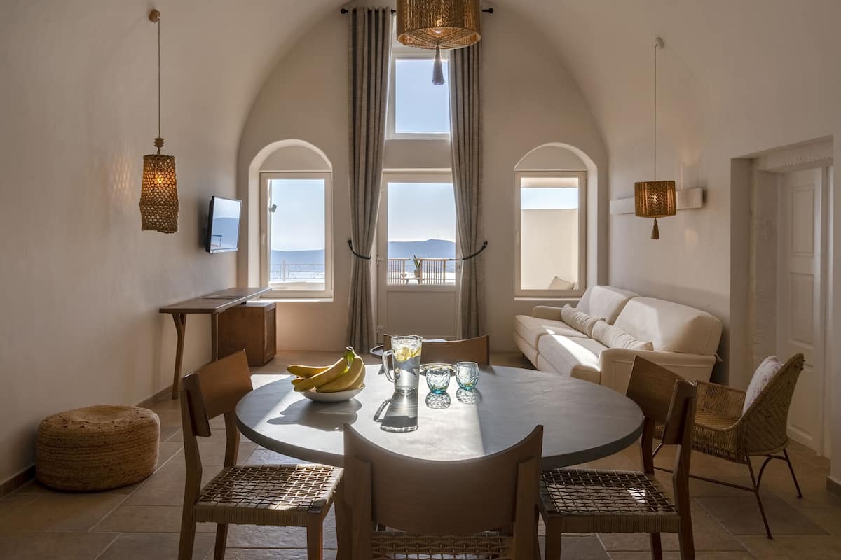 A dining area with a round table, chairs, a bowl of fruit, a pitcher, and glasses. There are two hanging lamps, a television, and a view of the sea through large windows.