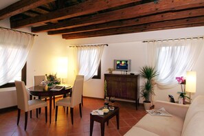 A living room with a sofa, two chairs, a coffee table, a TV, and a dining table with a vase of flowers. - Residence Corte Grimani (Venice)