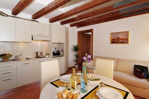 A dining area with a table set for two, a kitchen in the background, and a sofa with a painting on the wall. - Residence Corte Grimani (Venice)