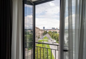 A view from a window with a balcony, overlooking a city street with buildings, greenery, and a clear sky.
