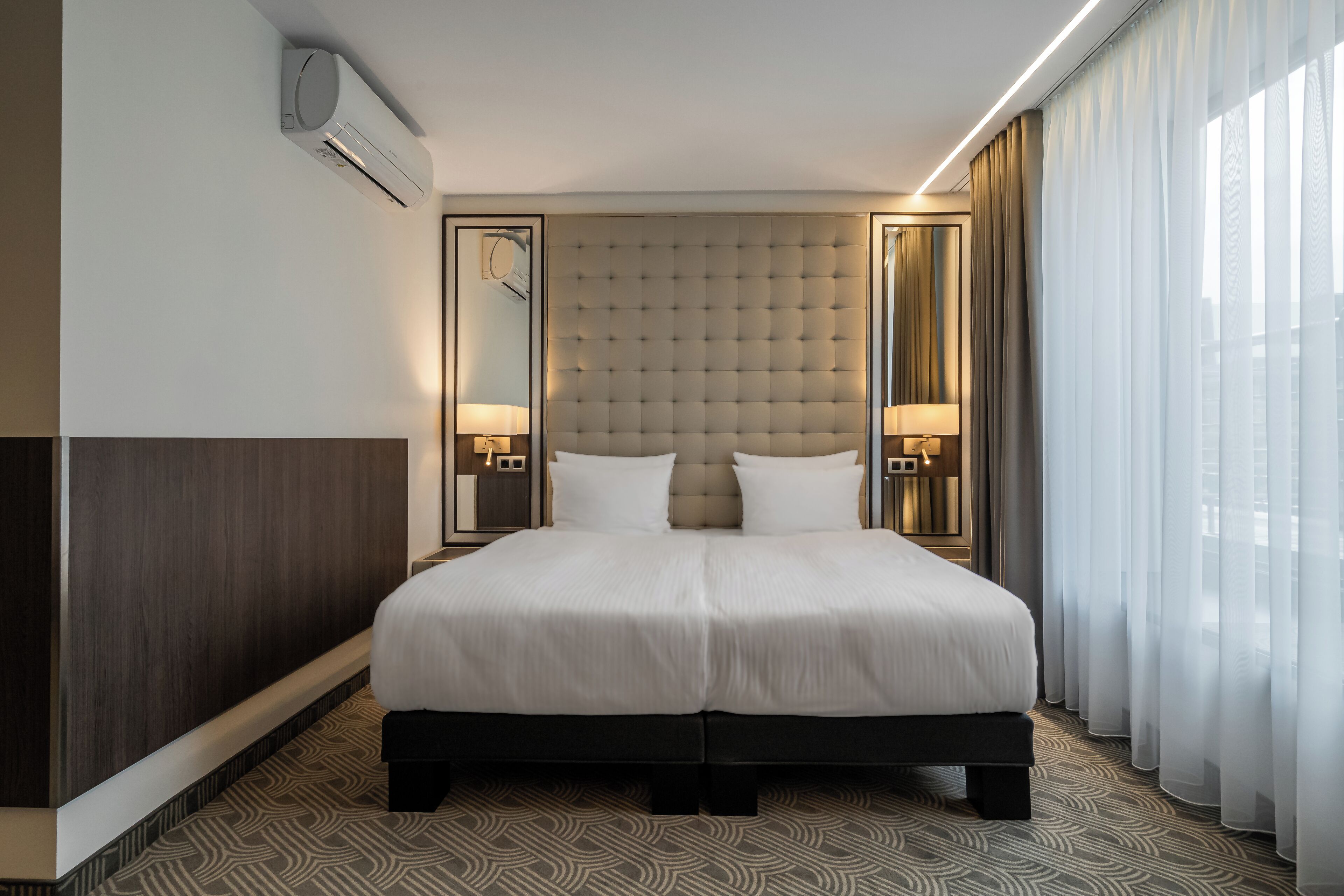 Una habitación de hotel moderna con una cama grande, dos lámparas de noche, un aire acondicionado montado en la pared y un ventanal con cortinas translúcidas.