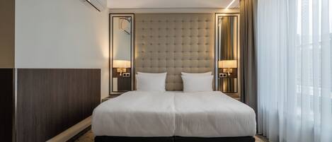 A modern hotel room with a large bed, two bedside lamps, a wall-mounted air conditioner, and a large window with sheer curtains.
