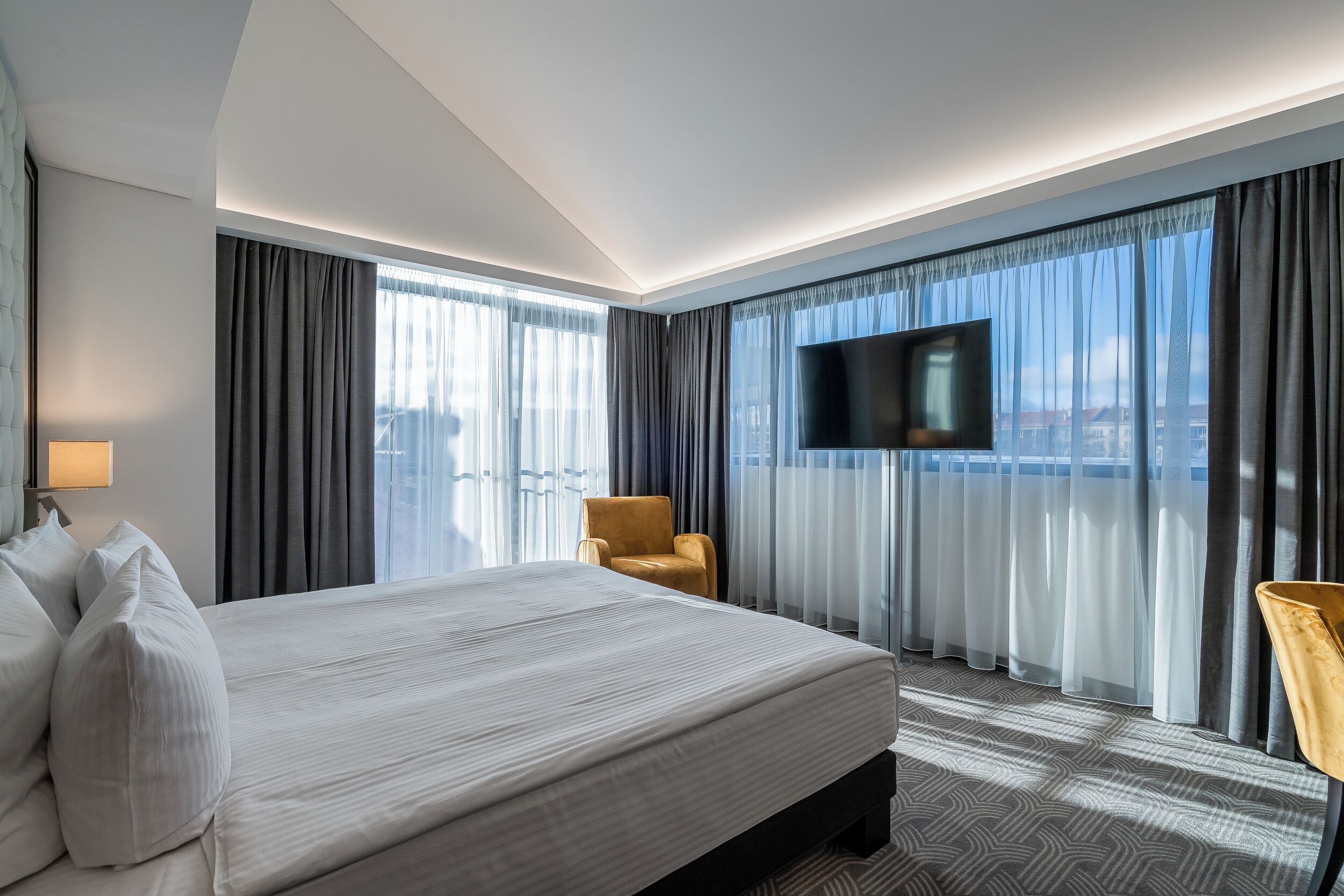 A modern hotel room with a large bed, a flat-screen TV, a comfortable armchair, and a city view through large windows.