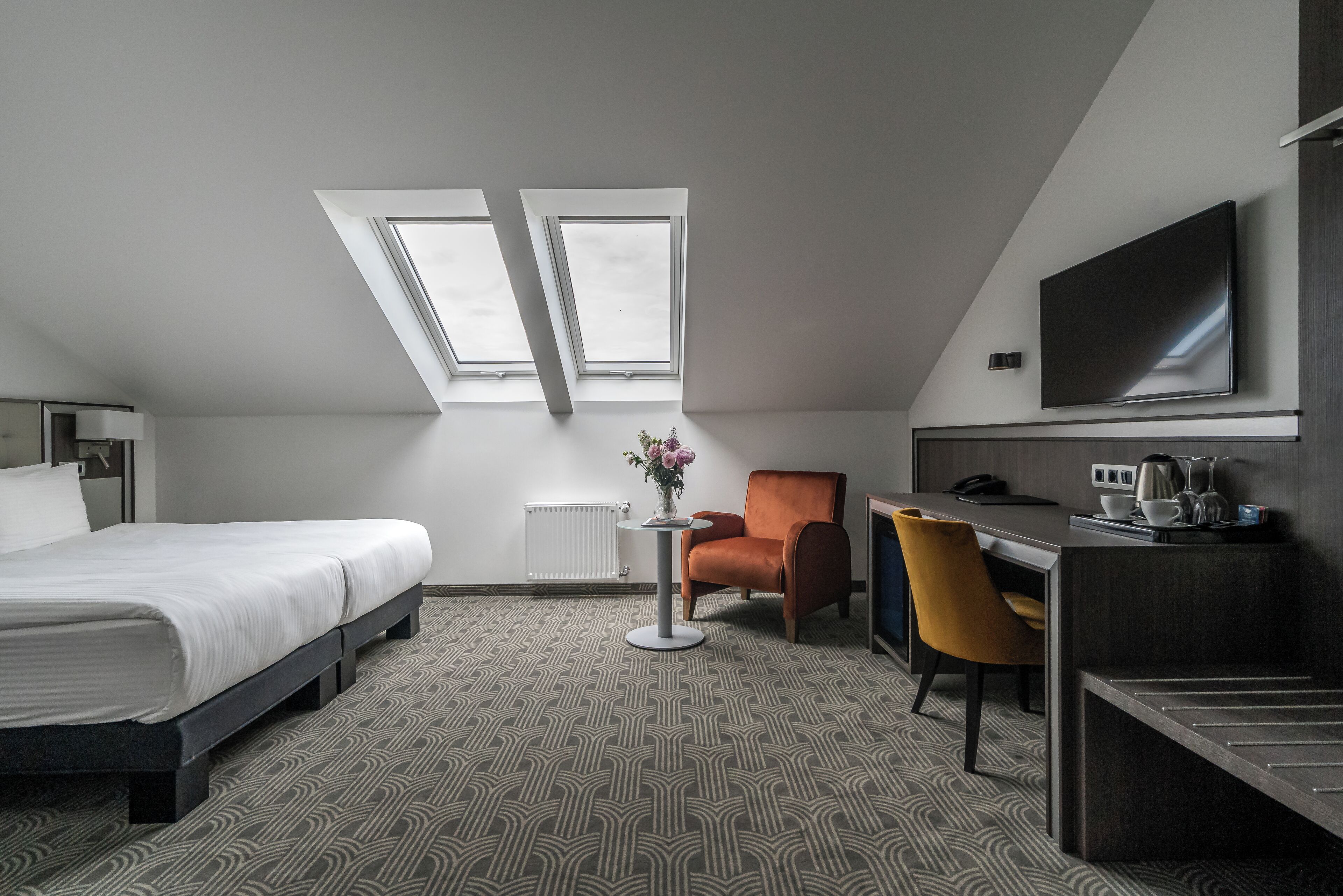 A hotel room with a bed, a desk, a chair, a TV, and two skylights.