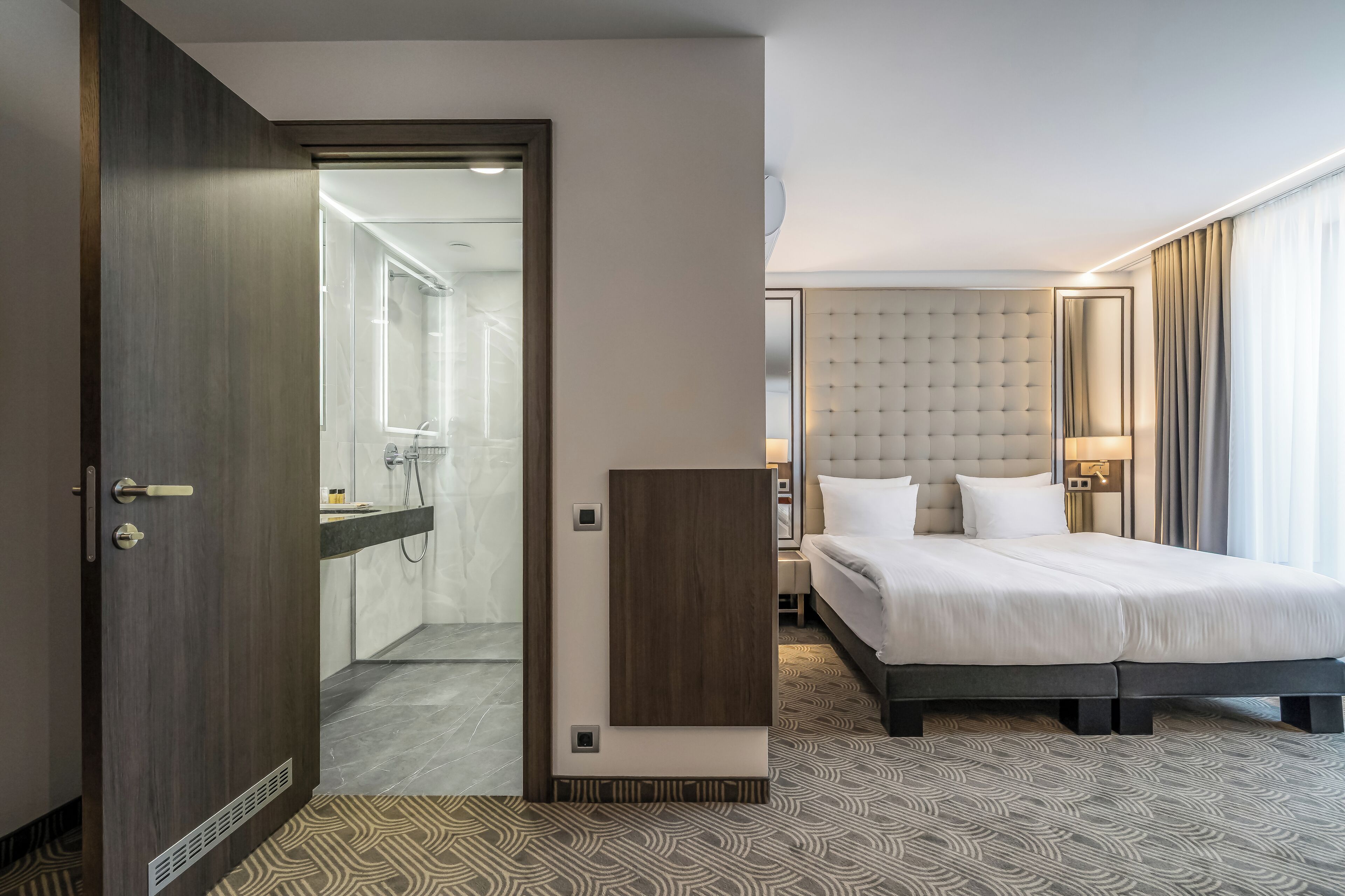 A hotel room with a bed, a bedside table, a mirror, a wardrobe, and a bathroom visible through an open door.