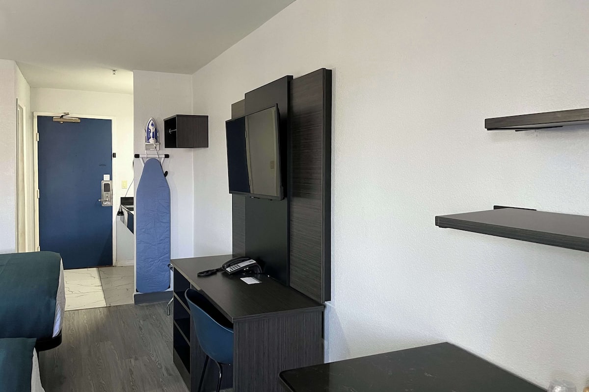 Una habitación de hotel moderna con un escritorio de madera oscura, una silla azul, un televisor de pantalla plana montado en la pared y un estante pequeño.