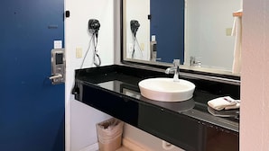 A bathroom with a black countertop, white sink, and a mirror. There is a trash can and a towel on the counter. - Studio 6 Suites Delano, CA (Delano)