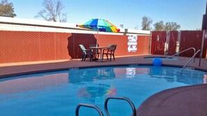 Une piscine entourée d’une clôture, une table avec des chaises et un parasol coloré.