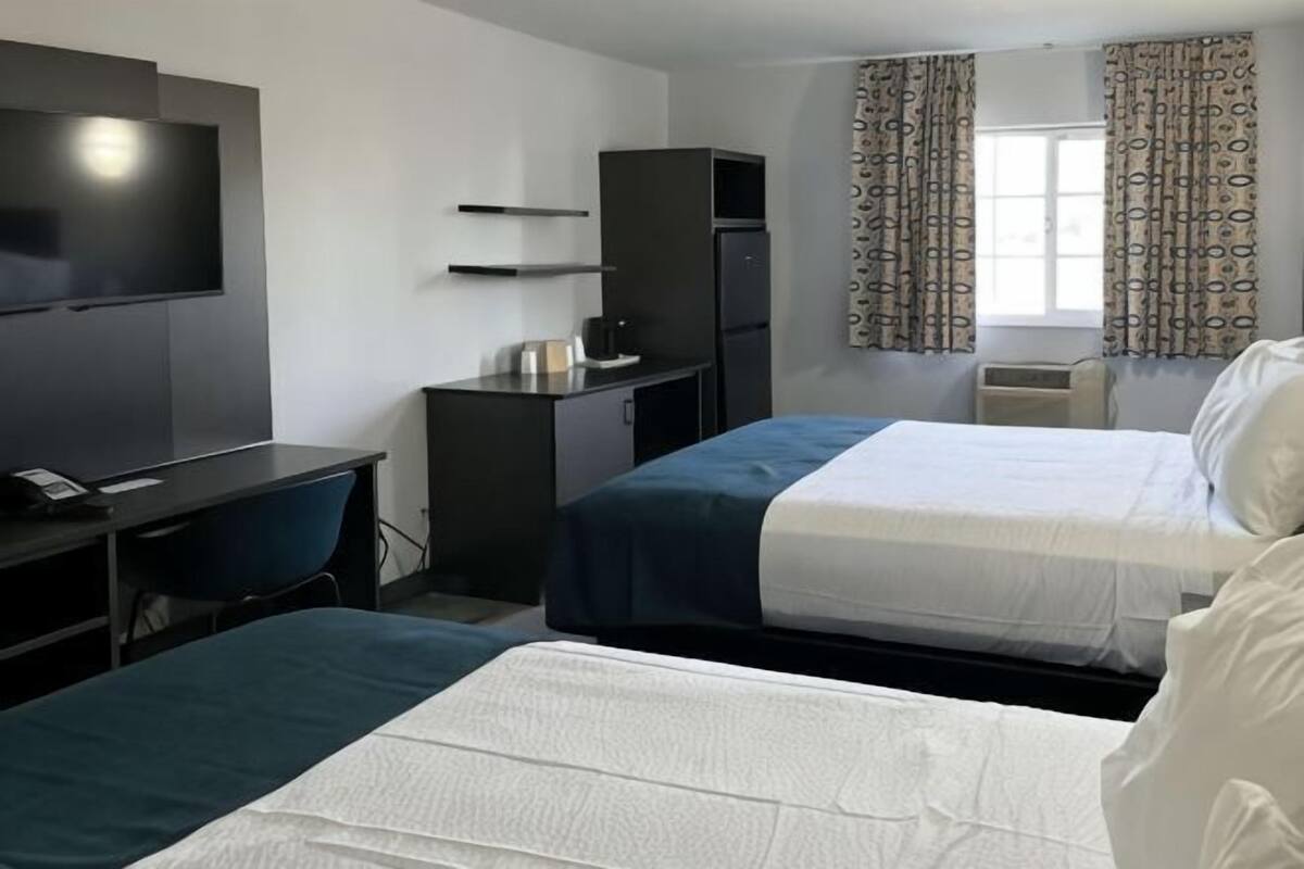 Habitación de hotel con dos camas, un escritorio con silla, televisión, refrigerador y ventana con cortinas estampadas.
