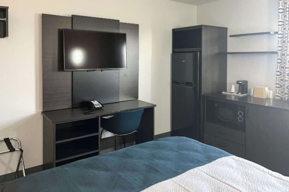 A hotel room with a bed, a desk with a chair, a flat-screen TV mounted on the wall, a built-in refrigerator, and a microwave.