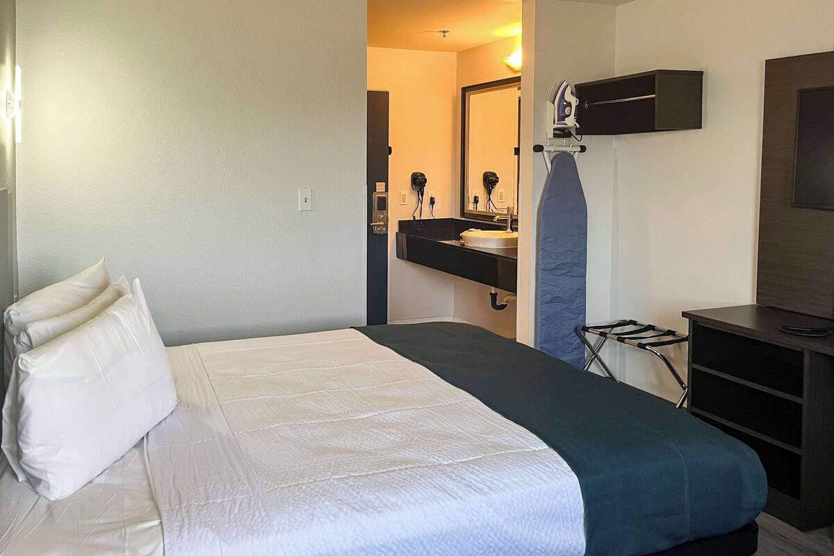 Habitación de hotel con cama, escritorio, espejo y baño visible a través de una puerta abierta.