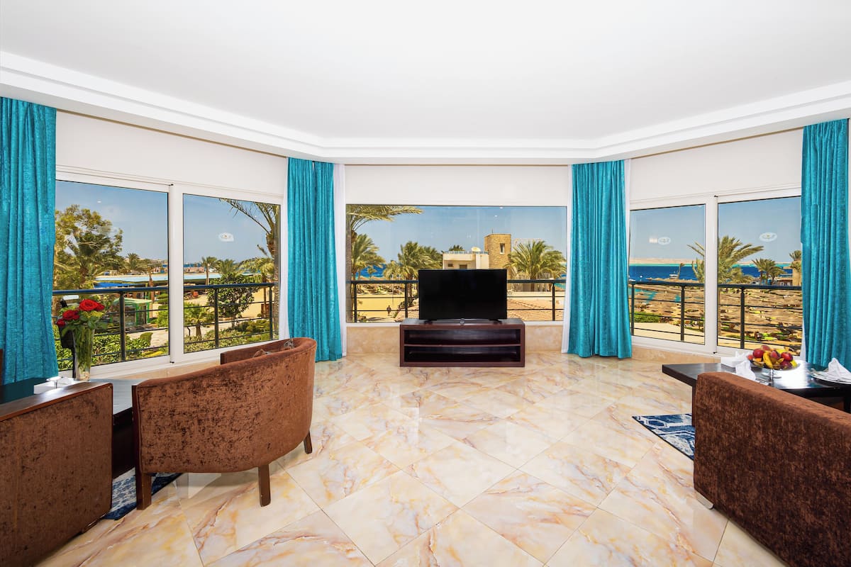 A spacious living area with a large TV, comfortable seating, and a balcony with a view of palm trees and buildings.