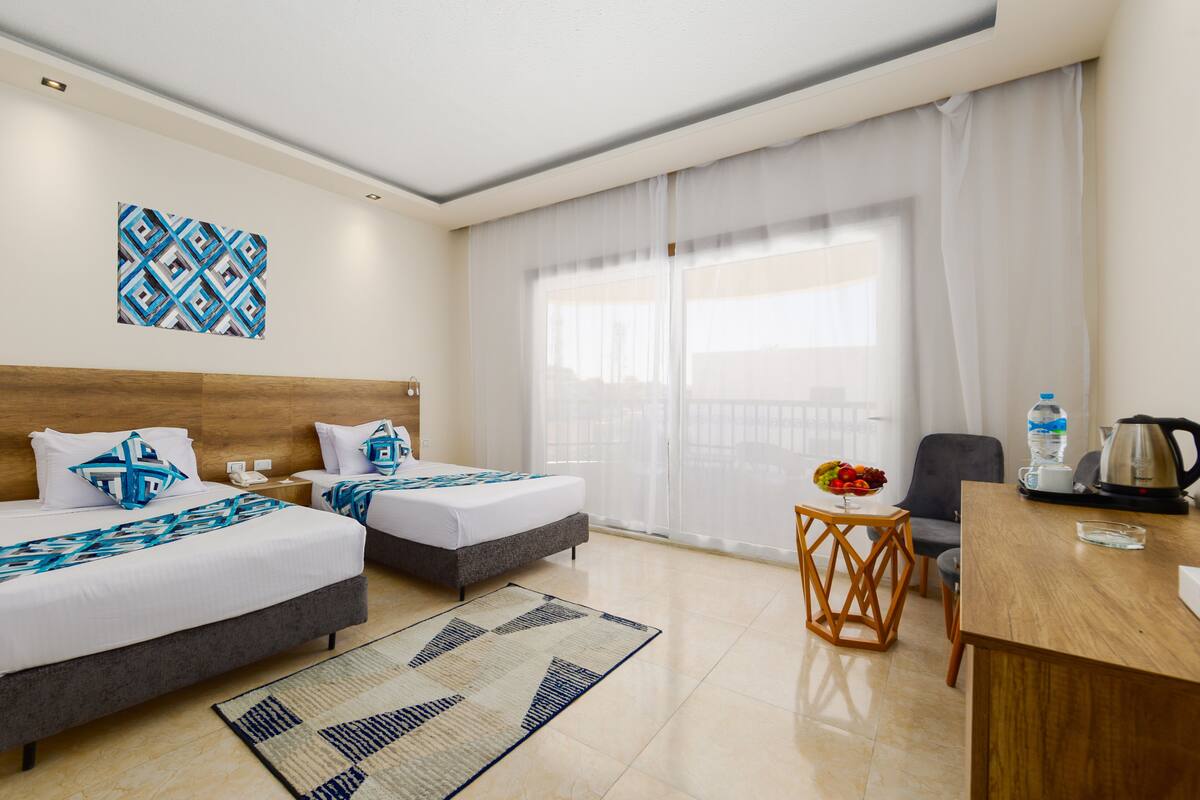 A hotel room with two beds, a wooden headboard, a small table with a fruit bowl, a coffee maker, and a view of the city through sheer curtains.