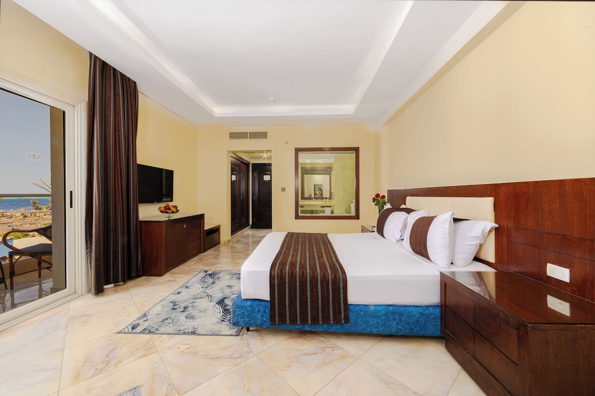 A hotel room with a bed, a TV, a desk, and a view of the beach.