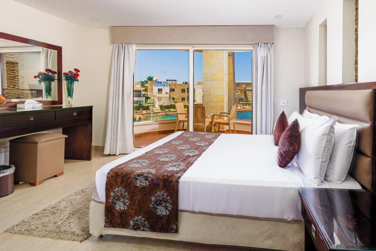 A hotel room with a large bed, a desk, a chair, and a view of a pool and buildings.