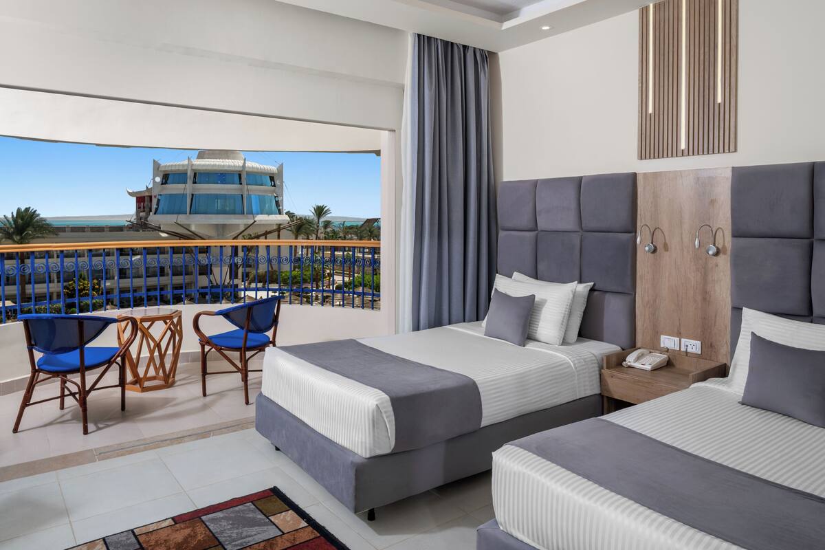 A hotel room with two beds, a balcony with a view of a building, and a table with chairs.