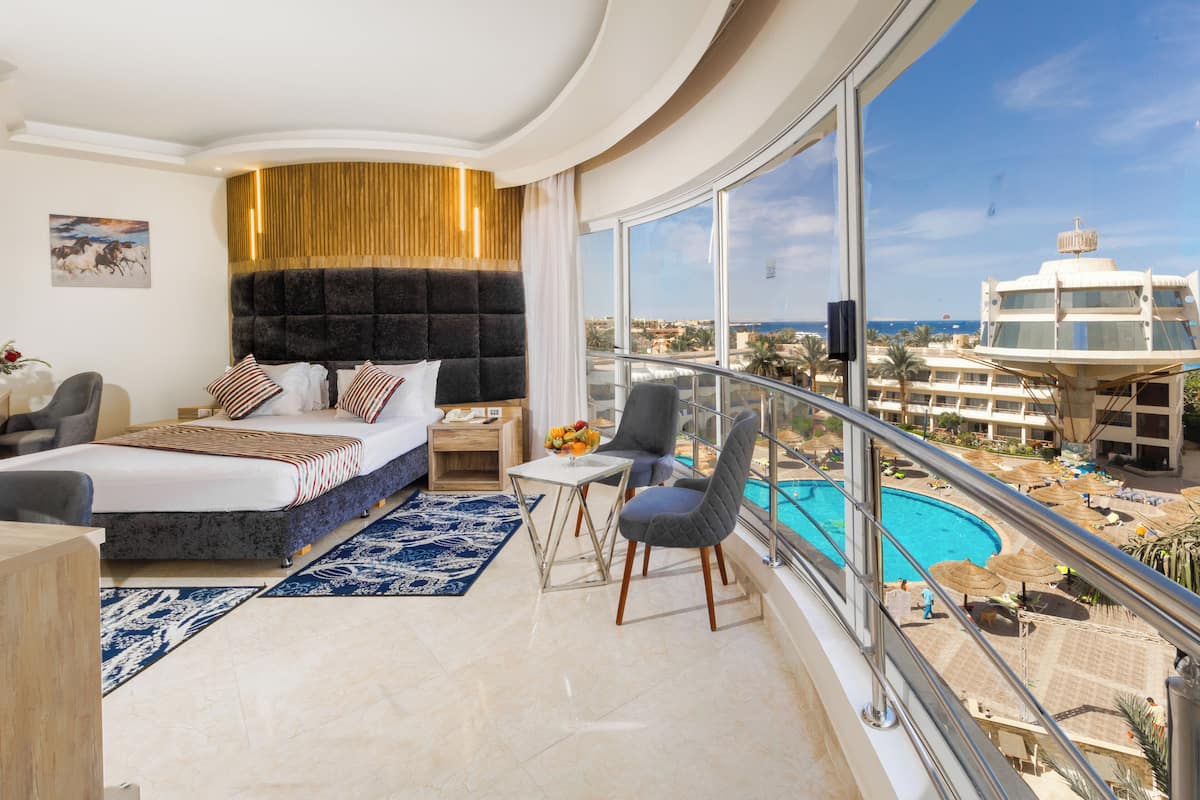 A modern hotel room with a large bed, a small table, a chair, and a balcony with a pool view.