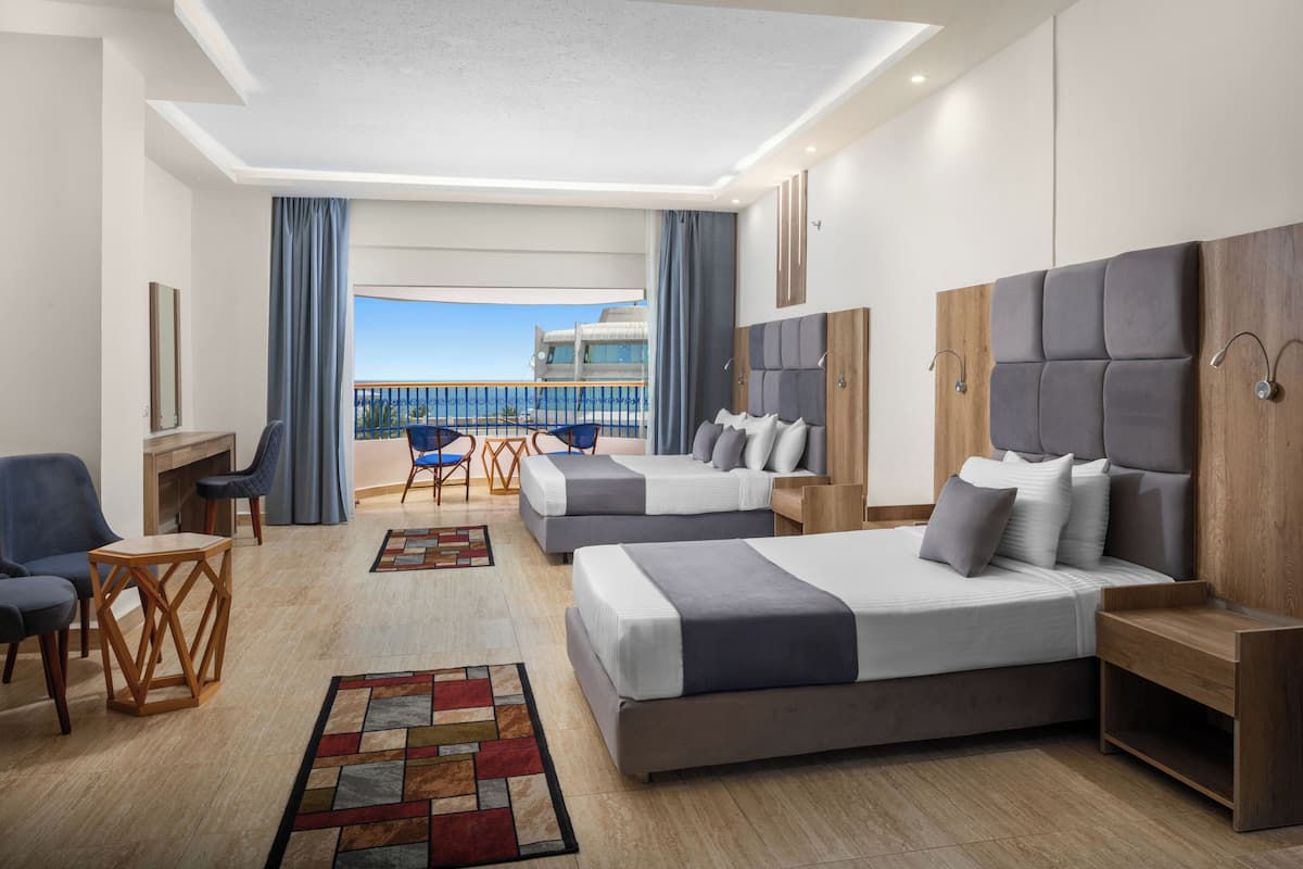 A hotel room with two beds, a desk, a chair, and a view of the sea.
