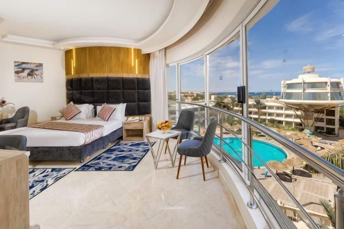 A modern hotel room with a curved ceiling, a large bed, a balcony with a view of the city and pool, and a small table with chairs.