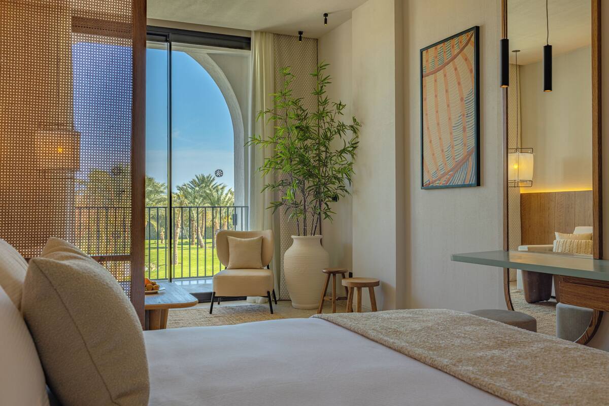 A hotel room with a large bed, a chair, a small table, a potted plant, and a balcony with a view of greenery.