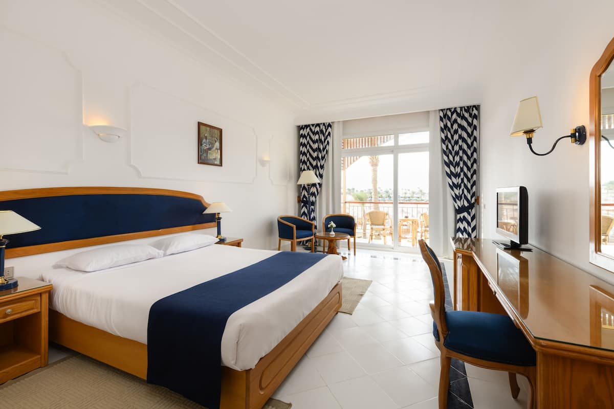 A hotel room with a large bed, wooden headboard, a desk with a chair, a television, and a balcony with outdoor seating.