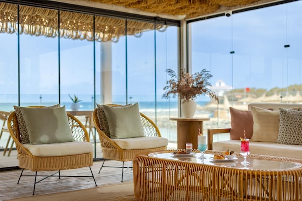 Un salon de plage aménagé avec des meubles en osier, une table ronde avec des boissons, et une vue sur l’océan.