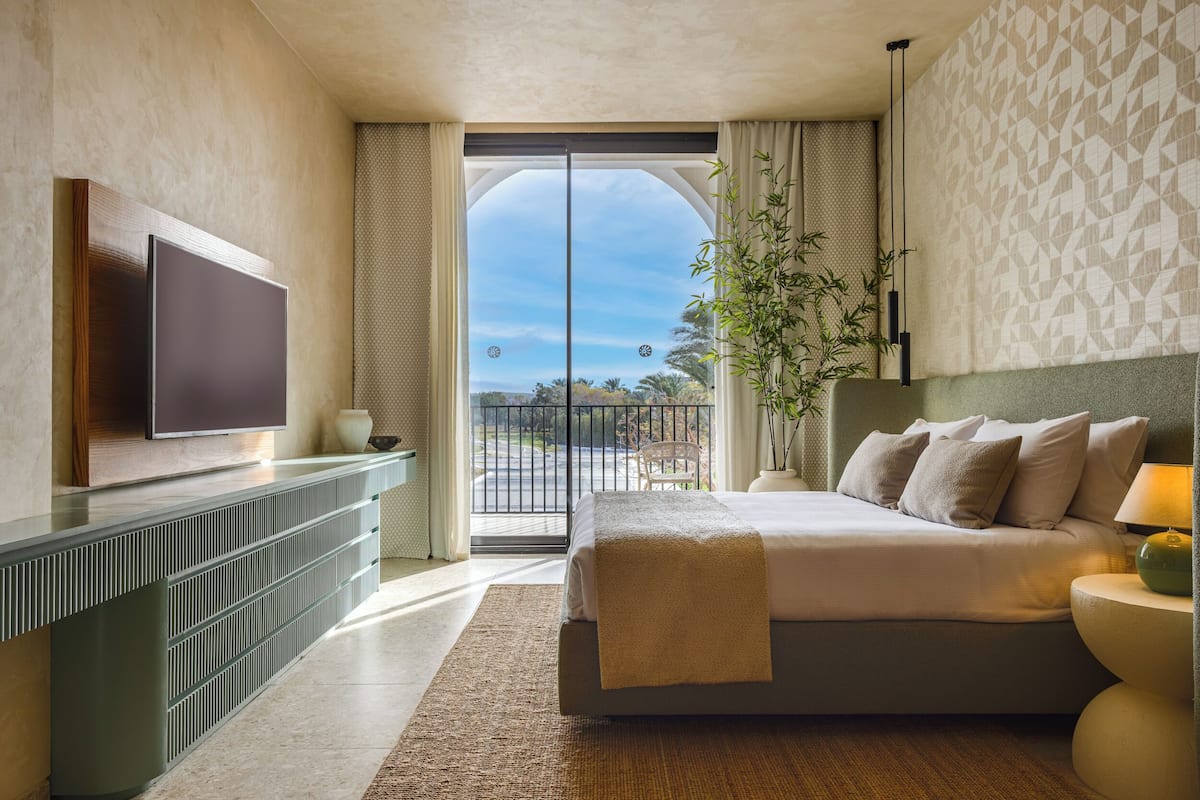 A modern hotel room with a large bed, a flat-screen TV, a balcony with a view, and a potted plant.