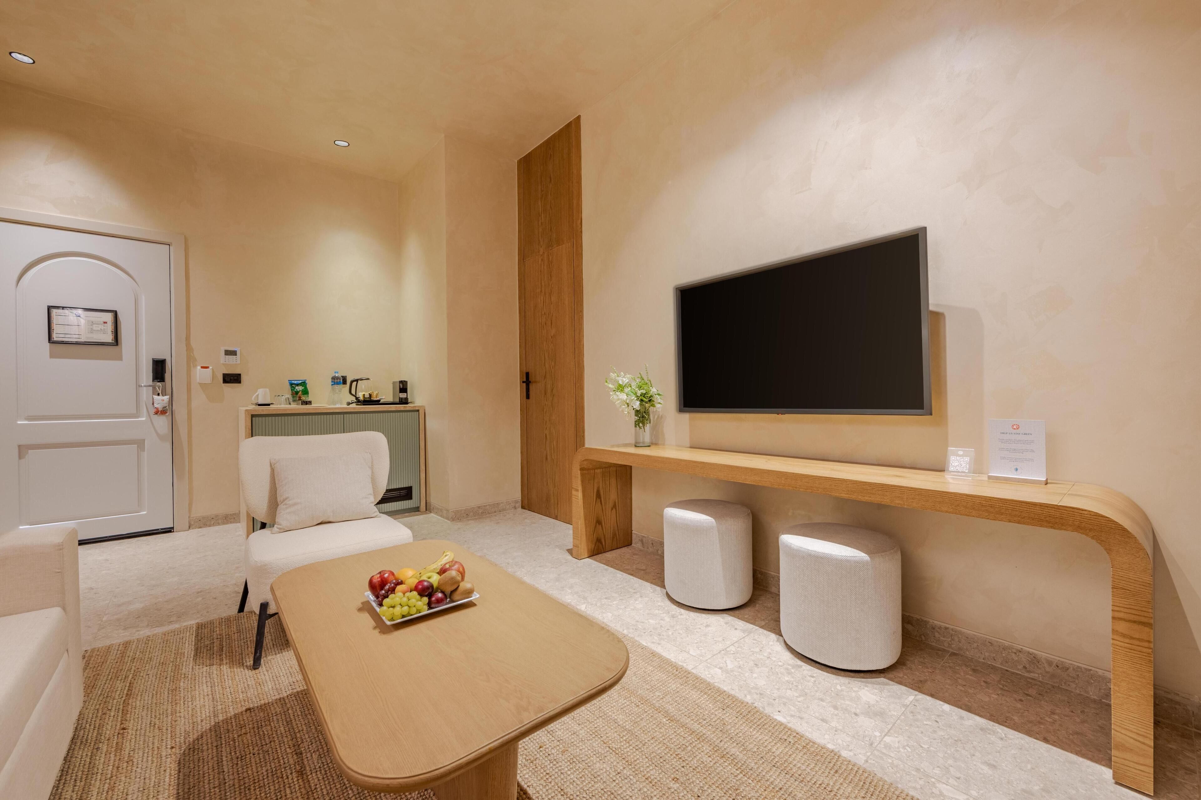 A modern hotel room with a wooden reception desk, a flat-screen TV, a seating area with a coffee table, and a small plant.