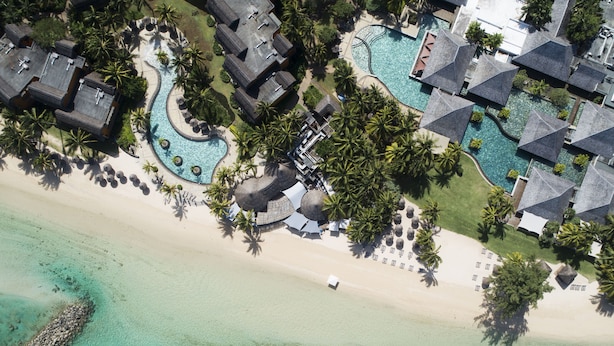 Vista aérea de un resort con múltiples bungalows sobre el agua, un área de piscina central y una playa.