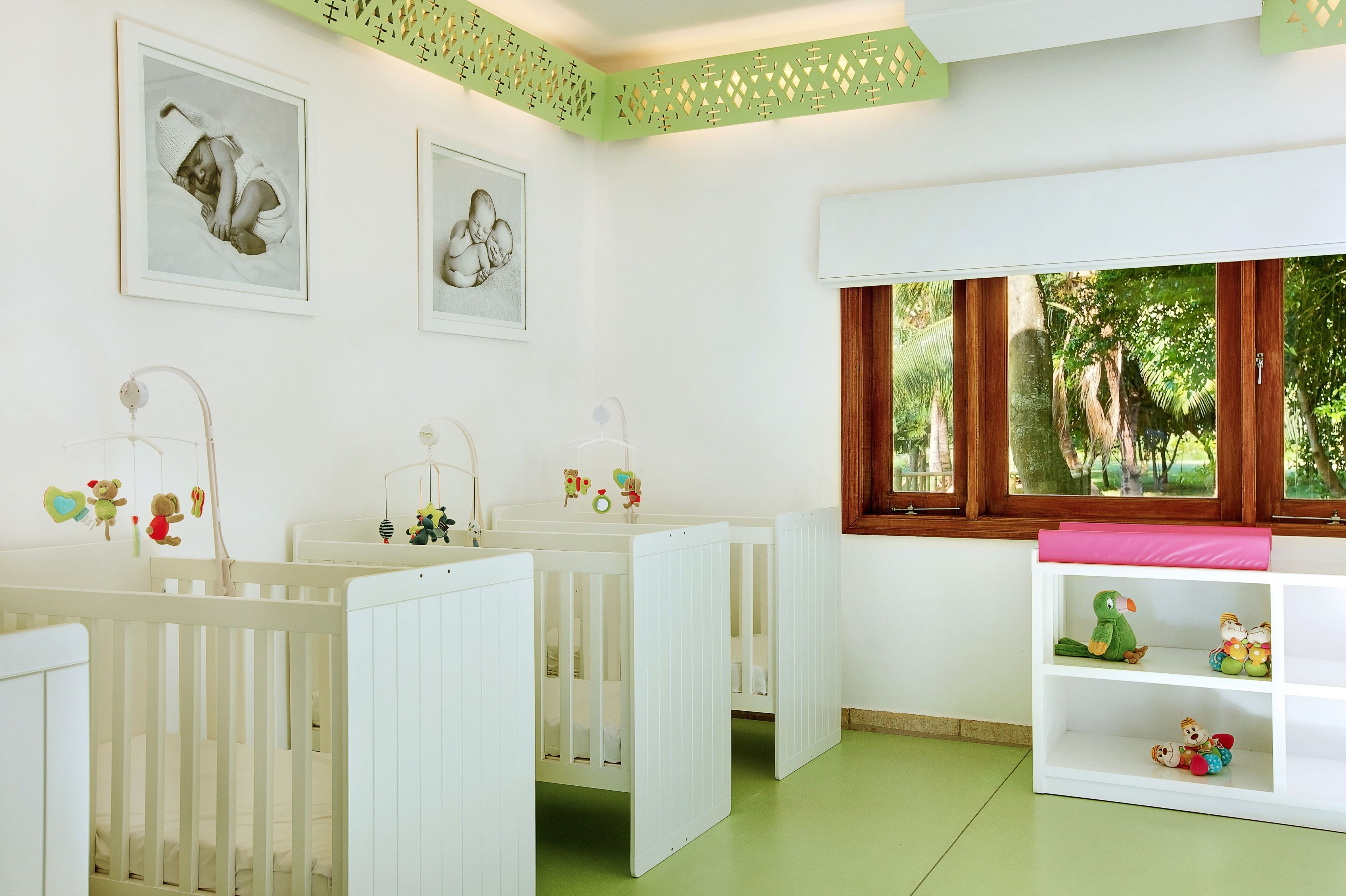 A baby room with two cribs, a window with a view of trees, and a white shelf with toys.