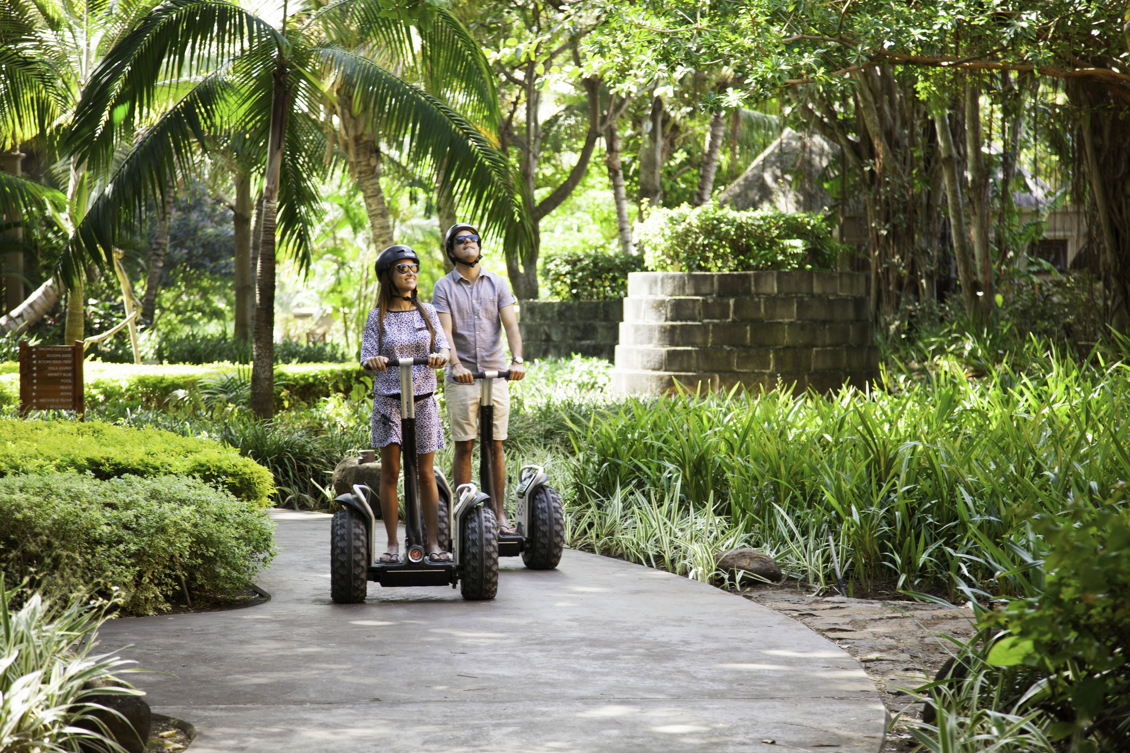 Two people on a paved path surrounded by lush greenery and palm trees.