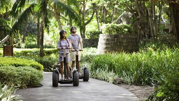 Two people on a paved path surrounded by lush greenery and palm trees.