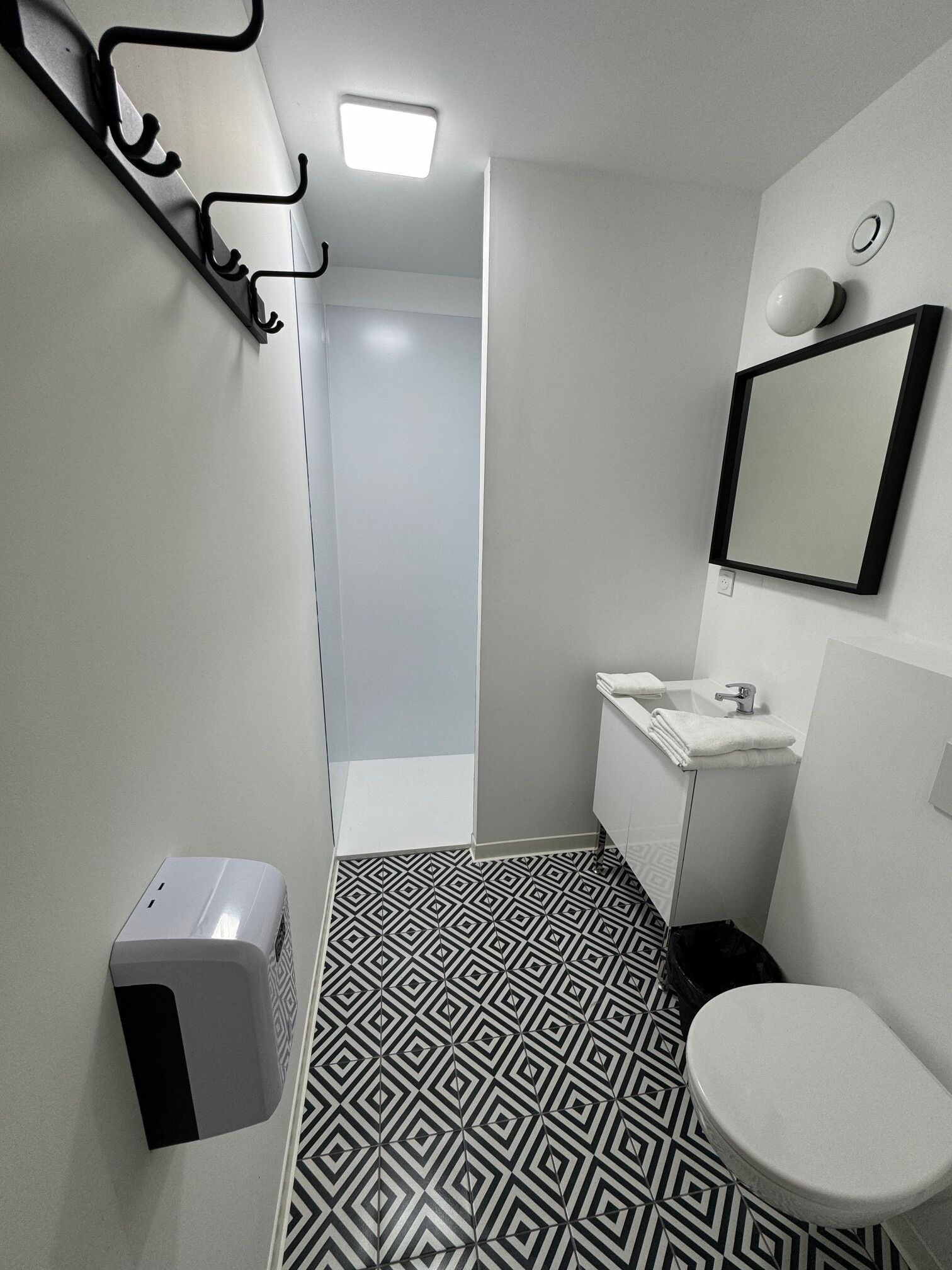 Ein modernes Badezimmer mit einem geometrischen Bodenmuster, einer weißen Toilette, einem Waschbecken mit Spiegel und einem Händetrockner.