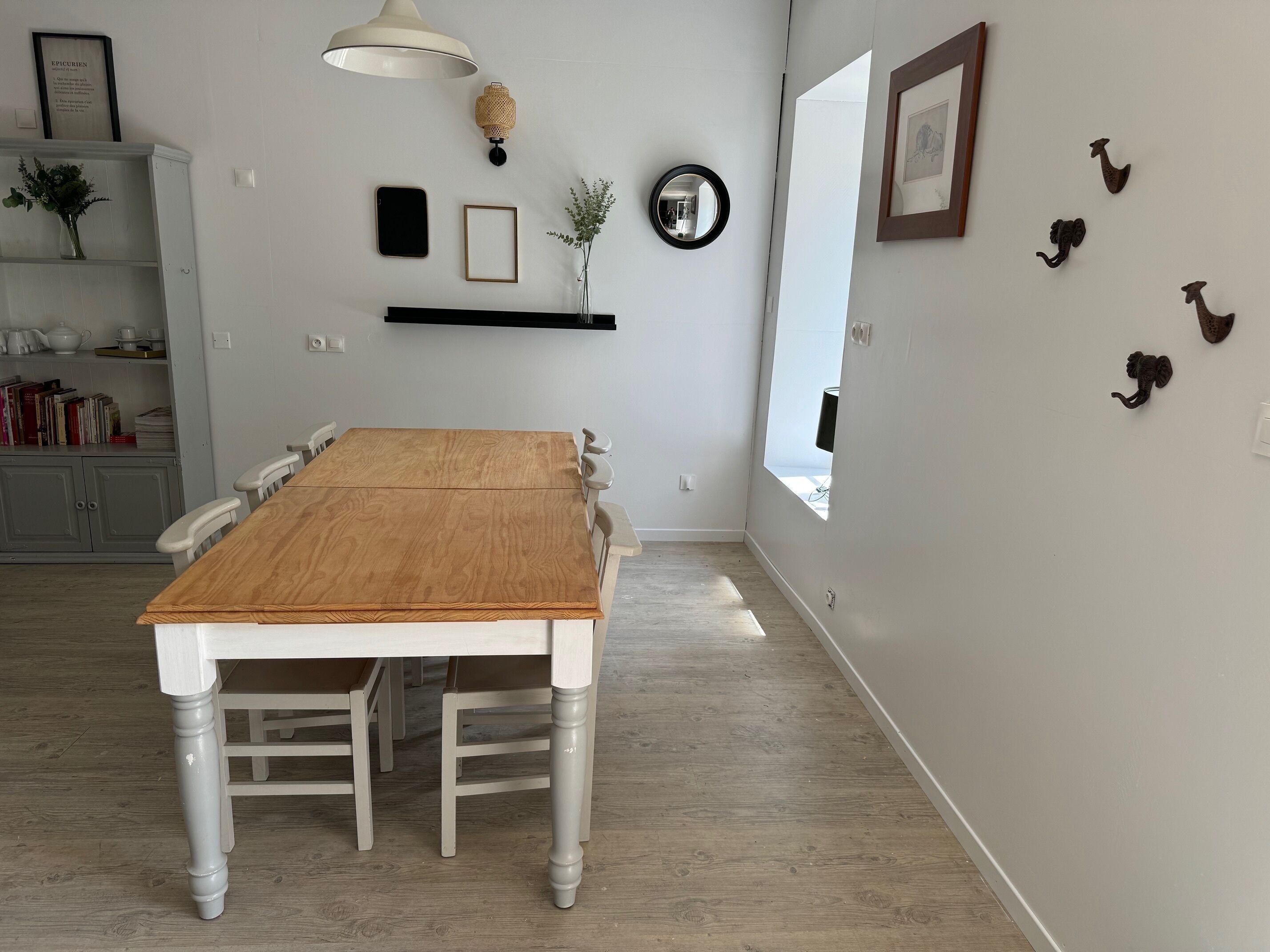 A dining area with a wooden table, white chairs, a white pendant light, a mirror, and wall-mounted decorations.