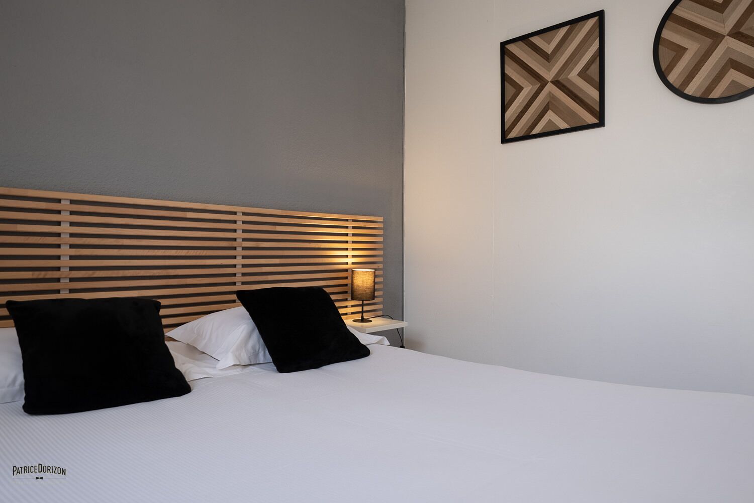 Una cama bien tendida con sábanas blancas y dos almohadas negras, un cabecero de listones de madera y una lámpara de noche.