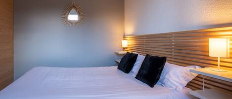 A hotel room with a bed, two bedside tables, a wall-mounted light fixture, and a wooden headboard.