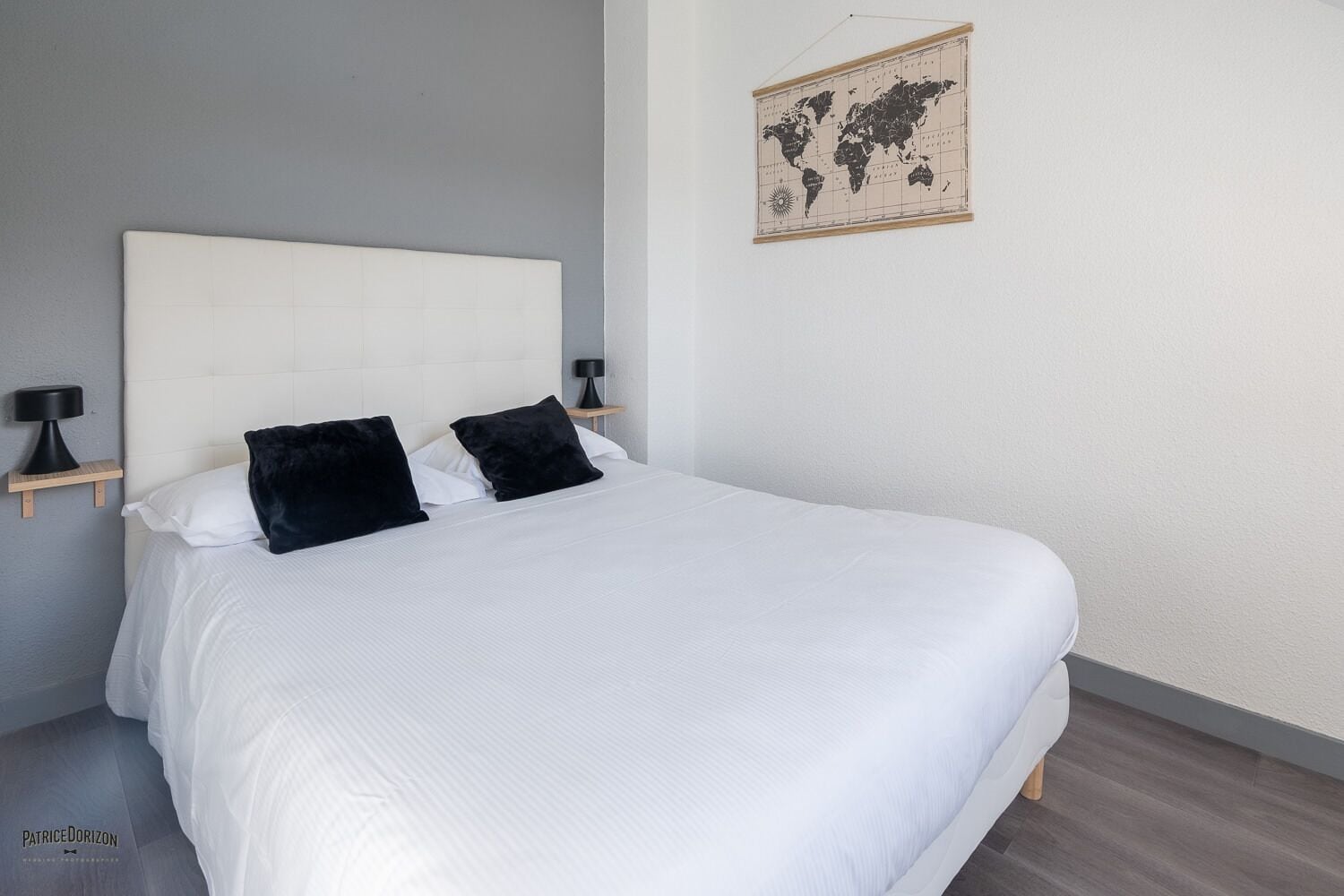Un letto rifatto con cura, con lenzuola bianche e due cuscini neri, una mappa del mondo appesa al muro e un comodino con una lampada accanto al letto.