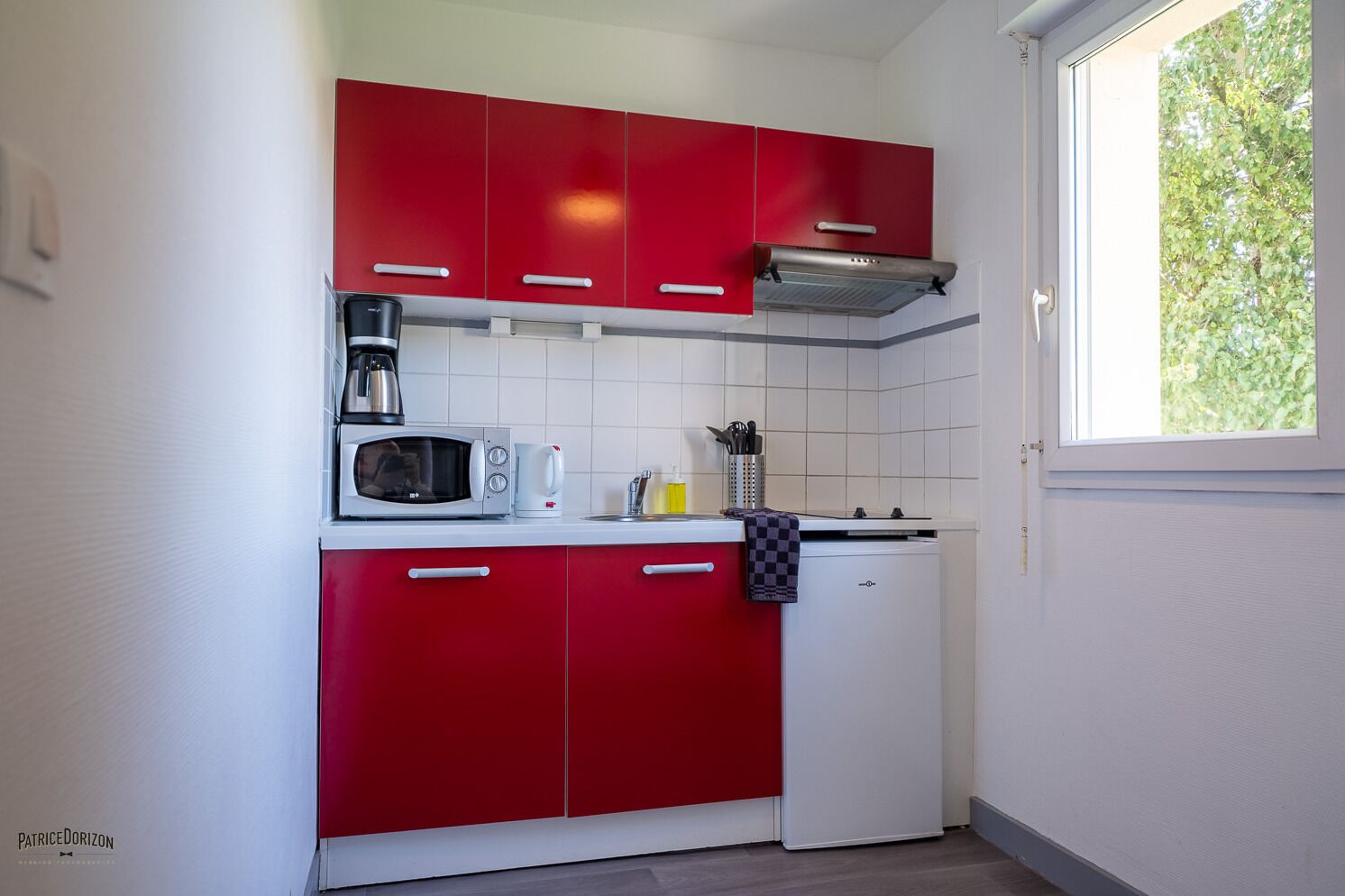 A compact kitchen with red cabinets, a microwave, a coffee maker, and a window with a view of greenery.